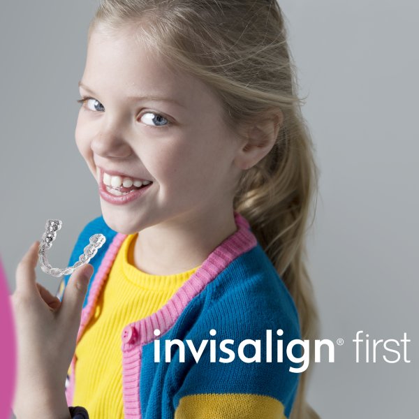 A smiling girl with her long hair tied in her back is holding an invisalign in her left hand and showing her left side view